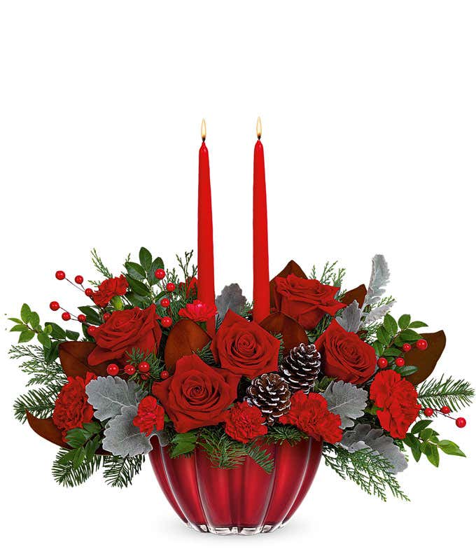 Red roses arranged with seasonal Christmas greens, pinecones, and berries in a shiny red bowl with two red tapered candles.