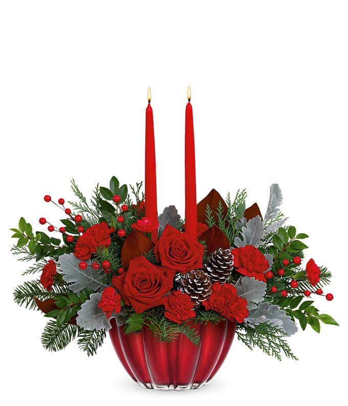 Red roses arranged with seasonal Christmas greens, pinecones, and berries in a shiny red bowl with two red tapered candles.