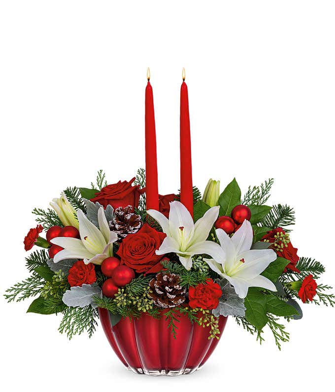 Roses and lilies arranged with Christmas greens, pinecones, and red ornament balls in a glassy red bowl with red tapered candles.