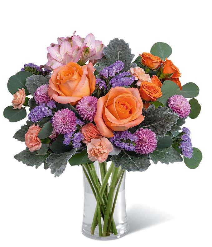 Short, round arrangement of orange roses and purple flowers, in a Tall Glass Cylinder Vase