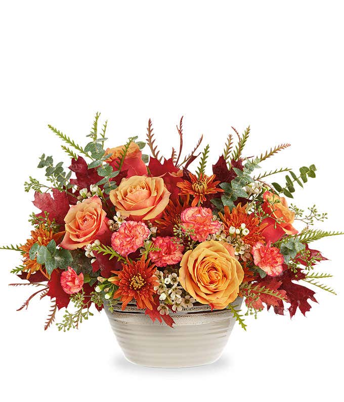 Light orange roses, orange carnations, bronze chrysanthemums, white waxflower, spiral & seeded eucalyptus, grevillea, and preserved red oak leaves in a rustic beige bowl against a white background