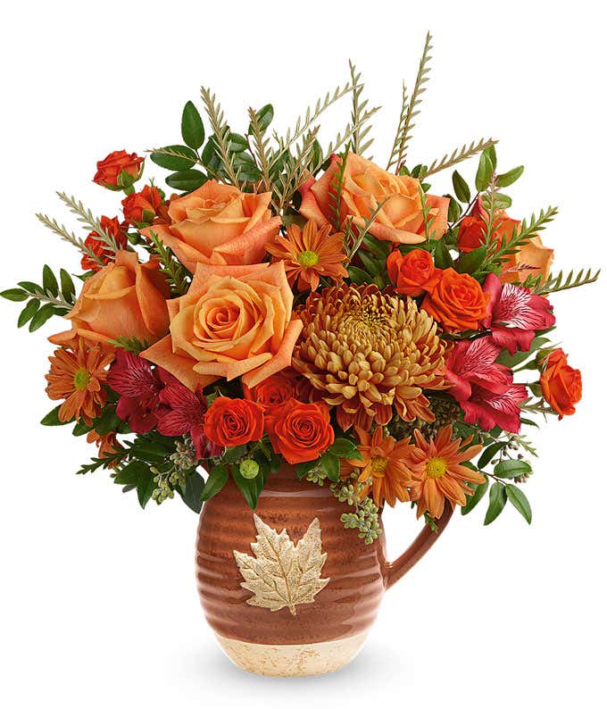 Orange roses, red alstromeria, orange spray chrysanthemums, bronze disbud chrysanthemums, seeded eucalyptus, in a light brown ceramic pitcher with a leaf against a white background