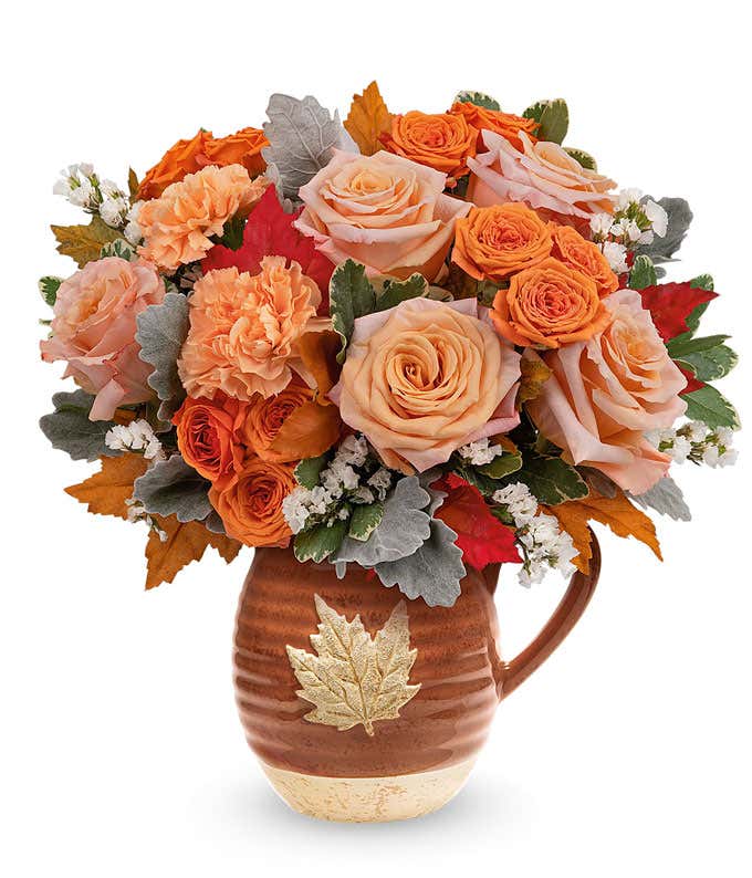 Peach and orange roses, orange carnations, white statice, variegated pittosporum, Red Oak Leaves in a brown ceramic pitcher against a white background