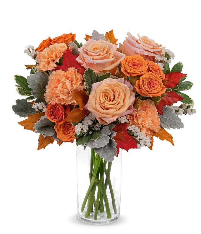 Peach and orange roses, orange carnations, white statice, variegated pittosporum, Red Oak Leaves in a brown ceramic pitcher against a white background