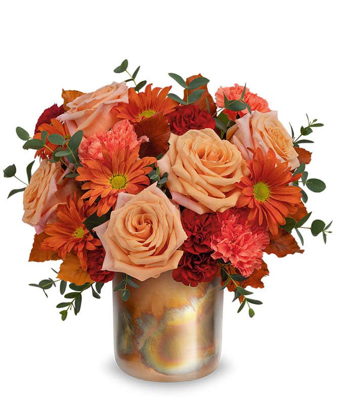 Light Orange Roses, Orange Carnations, Maroon mini carnations, bronze daisy spray chrysanthemums, brown copper beech, and parvifolia eucalyptus in a copper cylinder vase with patina against a white background