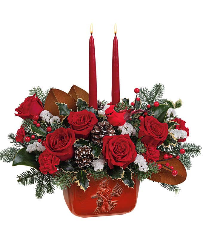 A festive arrangement with red roses, red miniature carnations, floral greens, a cardinal pick, red tapered candles, displayed in a keepsake red ceramic cardinal bowl.