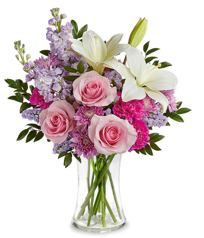 Image of a Mother's Day arrangement featuring Pink Roses, White Asiatic Lilies, Hot Pink Carnations, Lavender Stock, Lavender Cushion Spray Chrysanthemums, floral greenery, all beautifully presented in a metallic lavender vase.