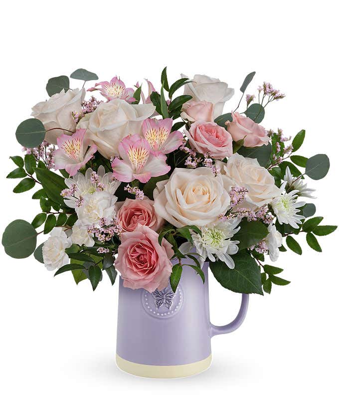 Image of a Mother's Day arrangement featuring Crme Roses, Pink Spray Roses, Light Pink Alstroemeria, White Cushion Spray Chrysanthemums, Pink Limonium, floral greens, all beautifully presented in a keepsake Lavender Butterfly Pitcher.
