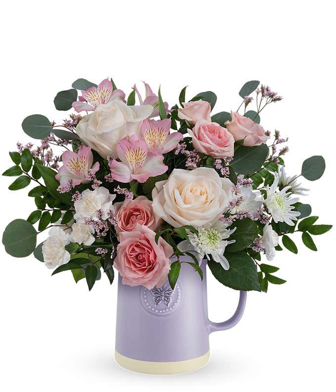 Image of a Mother's Day arrangement featuring Crème Roses, Pink Spray Roses, Light Pink Alstroemeria, White Cushion Spray Chrysanthemums, Pink Limonium, floral greens, all beautifully presented in a keepsake Lavender Butterfly Pitcher.