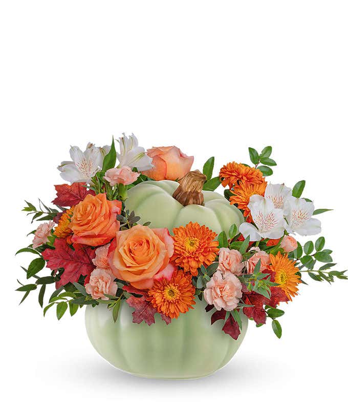 A fall floral arrangement featuring vibrant orange roses, white alstroemeria, miniature peach carnations, and bronze cushion spray mums, accented with seasonal greenery, all artfully arranged in a light green ceramic pumpkin-shaped container.