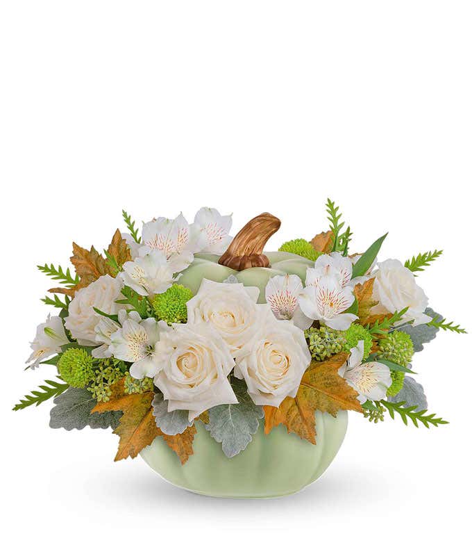 A delicate floral arrangement featuring white roses, white alstroemeria, and green button spray mums, accented with seasonal greenery, and presented in a light green ceramic pumpkin-shaped container with a brown stem accent.