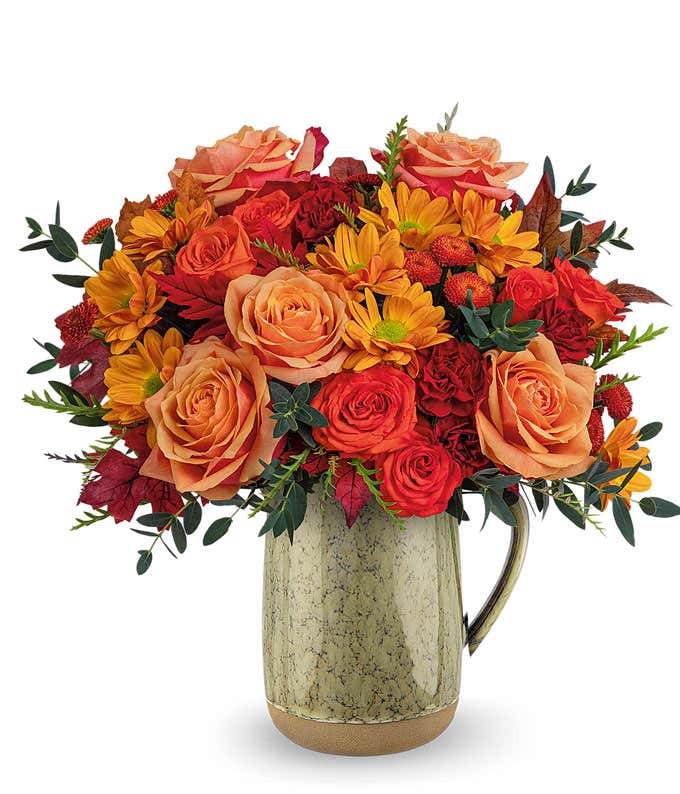 A warm floral arrangement featuring orange roses, orange spray roses, miniature maroon carnations, brown button spray mums, and orange daisy spray mums, accented with seasonal floral greenery, and presented in a rustic ceramic pitcher with a speckled fini