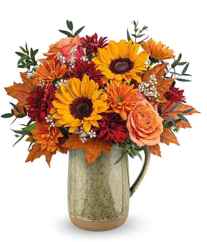 A cheerful floral arrangement featuring orange roses, red cushion spray mums, bronze daisy spray mums, and bright yellow sunflowers, accented with seasonal floral greenery, and presented in a rustic ceramic pitcher with a speckled finish.