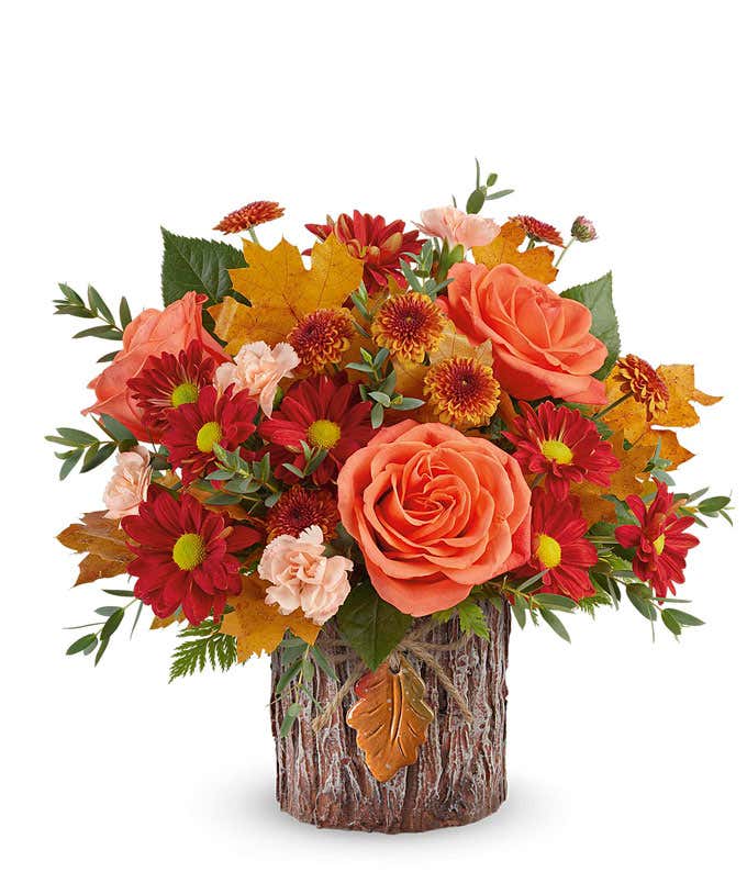 A vibrant arrangement with orange roses, miniature peach carnations, bronze button spray mums, red daisy spray mums, and seasonal greenery in a keepsake autumnal container with a rustic wood texture and leaf accent.