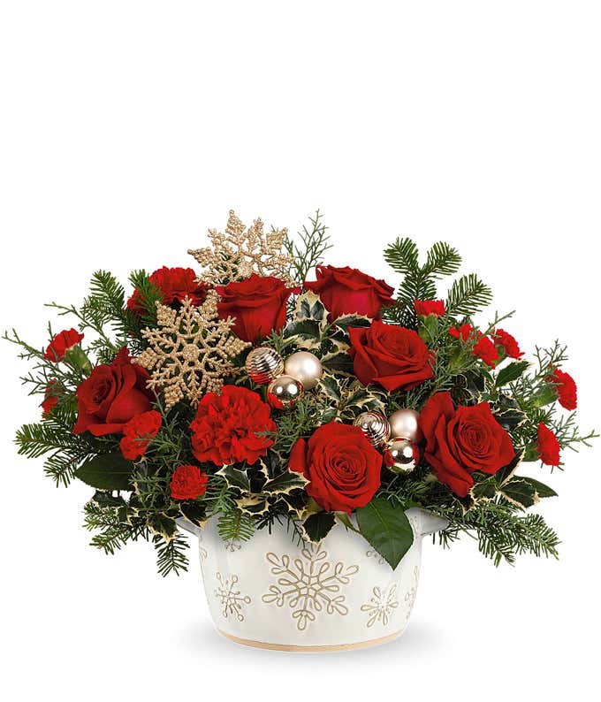 Holiday arrangement with red roses, red carnations, and seasonal greenery, accented by gold glitter snowflakes, gold ornaments, and evergreen sprigs. Presented in a white ceramic container with snowflake designs