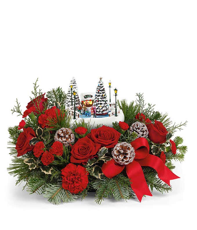 Festive holiday centerpiece featuring red roses, red carnations, white-tipped pinecones, holly, and evergreen sprigs. The arrangement is accented with a red bow and includes a miniature winter scene with snow-covered trees.