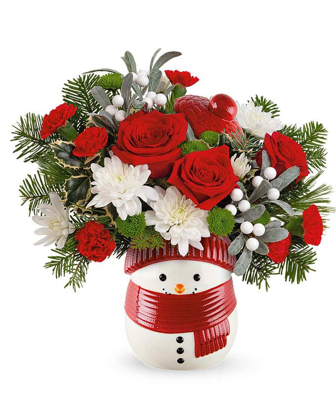 Festive holiday bouquet featuring red roses (deluxe & premium sizes), red carnations, white chrysanthemums, green button mums, and seasonal greenery, accented with white berries and arranged in a snowman-themed container with a red scarf and hat.