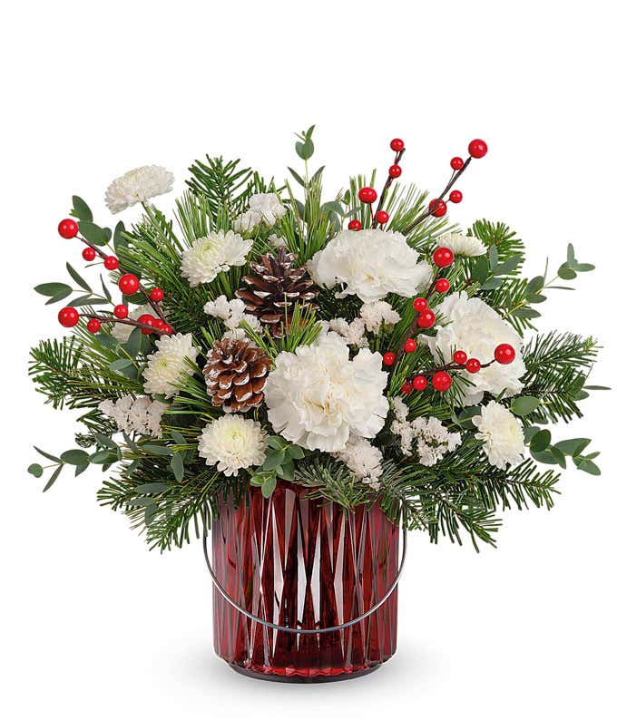 Holiday floral arrangement featuring white roses, white carnations, pinecones, red berries, and festive greenery, displayed in a red glass vase with silver accents, creating a wintery and elegant look.