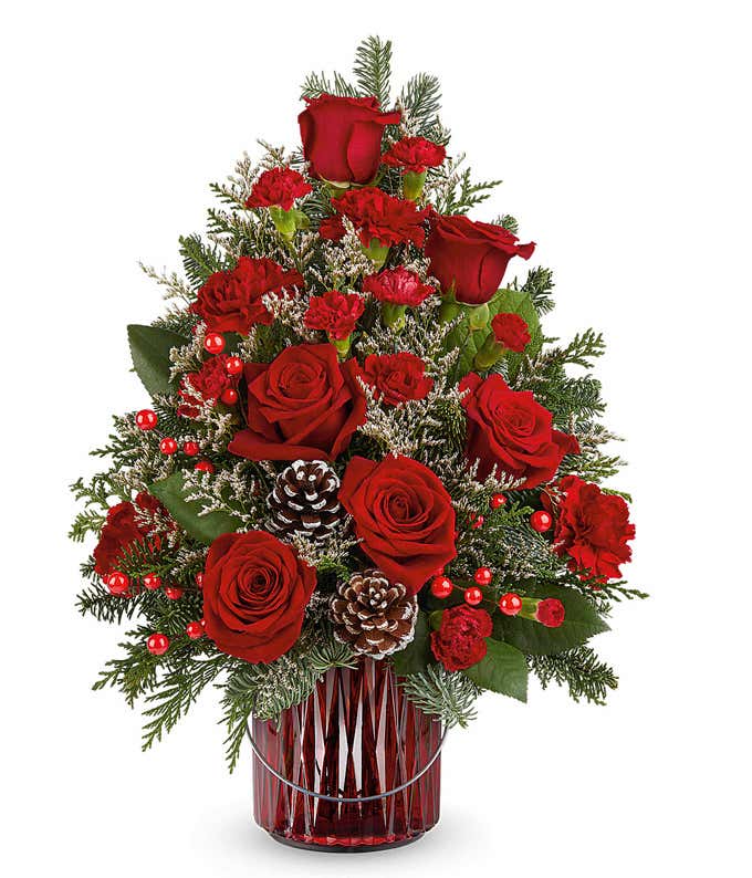 Christmas tree-shaped floral arrangement featuring red roses, red carnations, white-tipped pinecones, red berry accents, and evergreen sprigs in a red vase.