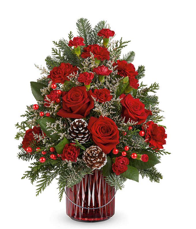 Christmas tree-shaped floral arrangement featuring red roses, red carnations, white-tipped pinecones, red berry accents, and evergreen sprigs in a red vase.