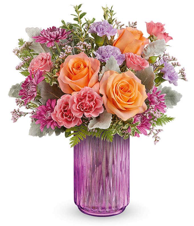 Vibrant floral arrangement featuring orange roses, pink miniature carnations, lavender carnations, pink cushion spray chrysanthemums, dusty miller, leatherleaf fern, and pink statice in a textured pink glass vase.