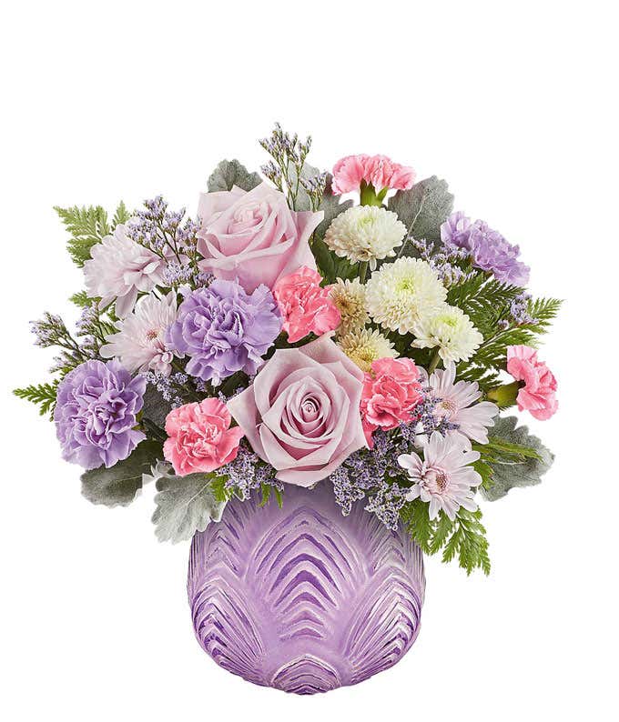 Soft pastel floral arrangement featuring lavender roses, lavender carnations, pink miniature carnations, lavender cushion spray chrysanthemums, white button spray chrysanthemums, dusty miller, and lush greenery in a textured lavender glass vase.