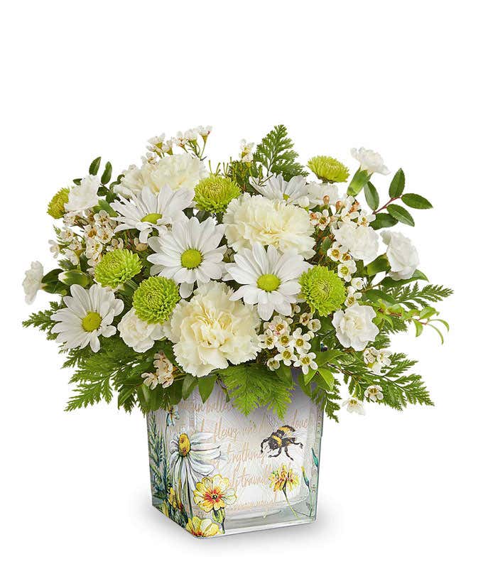 White roses, white daisies, white carnations, green button mums, white waxflower, and assorted greenery arranged in a square glass container with a watercolor-style bee and floral design.