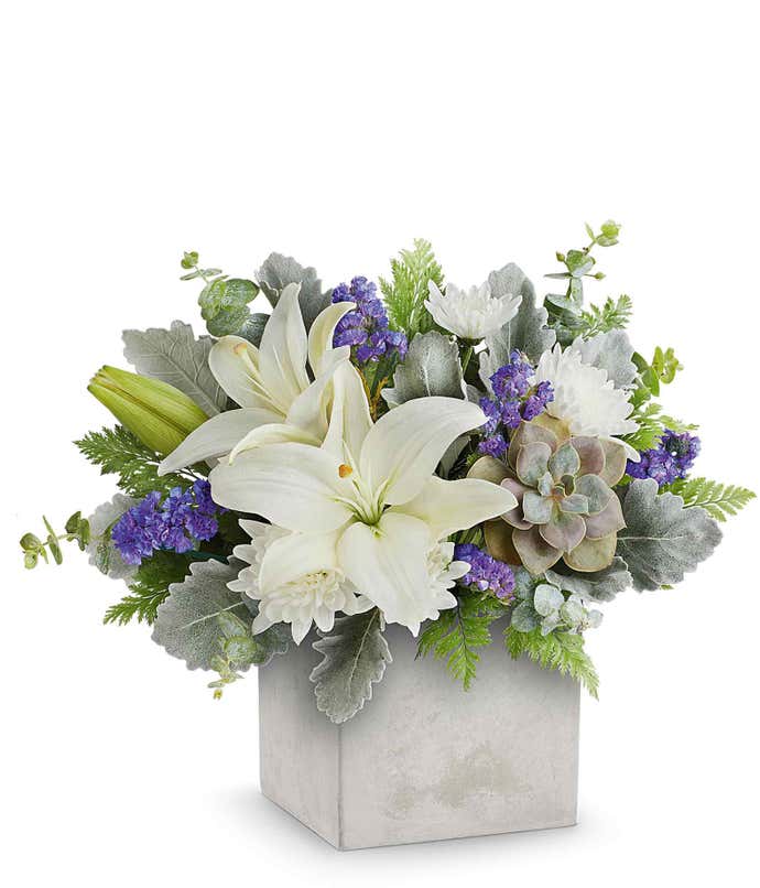A floral arrangement in a square gray container featuring white lilies, purple thistle, green succulents, silver foliage, and seasonal greenery.
