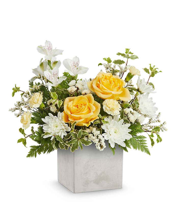 A floral arrangement in a square gray container featuring yellow roses, white alstroemeria, white carnations, white poms, ivy, and seasonal greenery.
