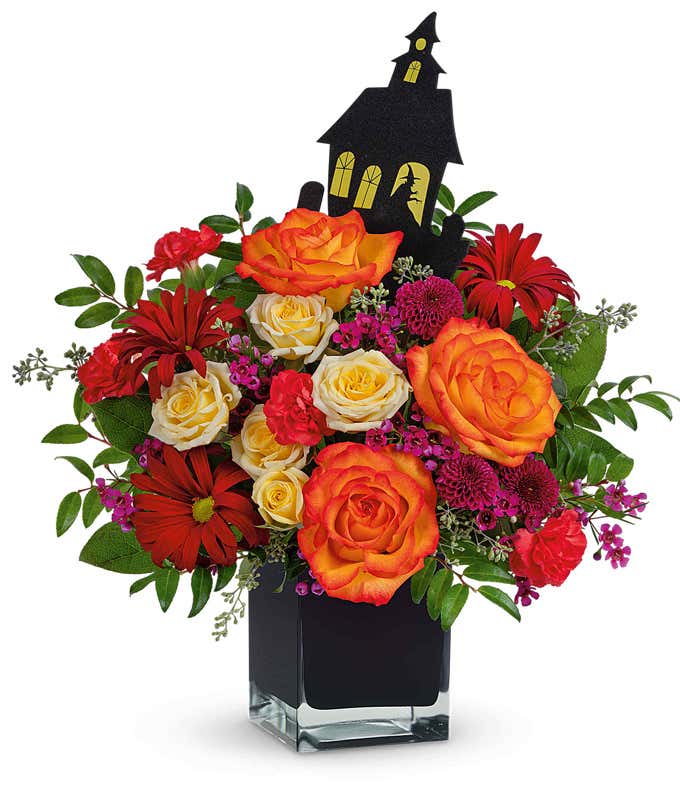 Halloween-themed flower arrangement with orange and yellow roses, red daisies, and pink filler flowers in a black cube vase, accented by greenery and topped with a black haunted house decoration.