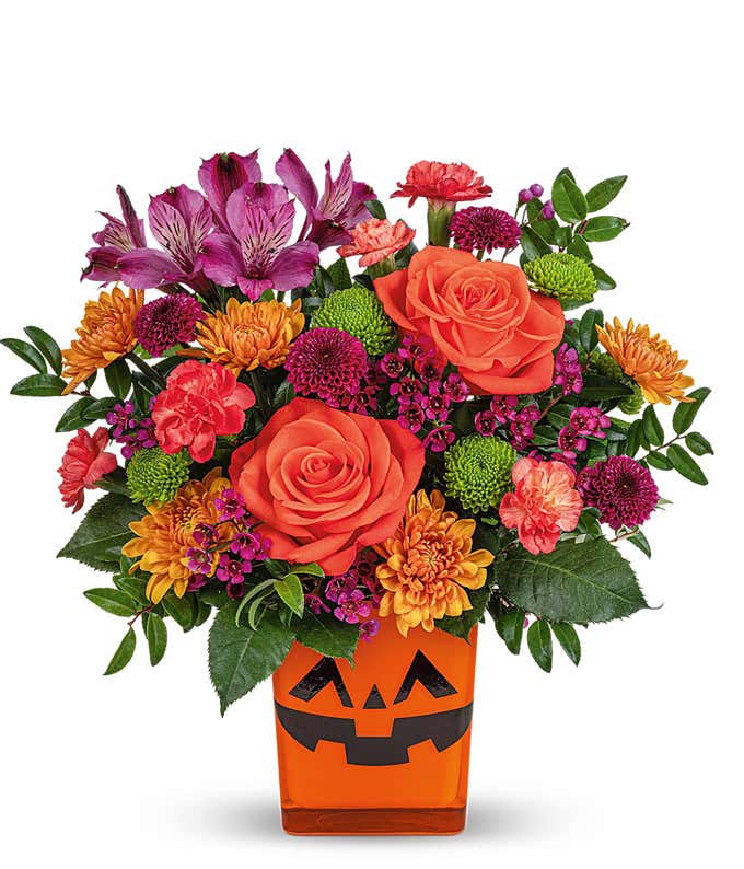 Halloween-themed flower arrangement featuring orange roses, purple alstroemeria, green button spray chrysanthemums, and assorted mums in a bright orange jack-o'-lantern container.