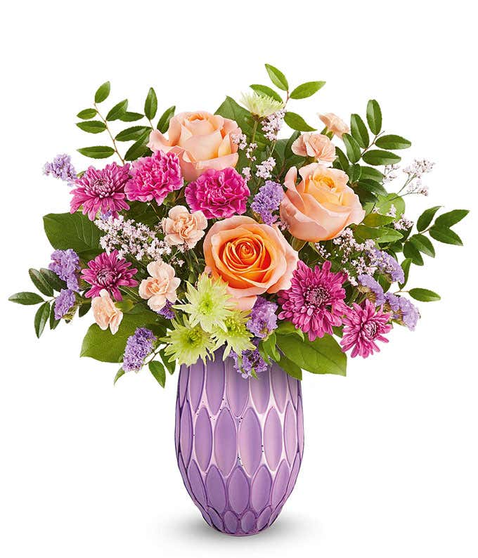 Peach roses, fuchsia carnations, miniature peach carnations, green cushion spray chrysanthemums, purple cushion spray chrysanthemums, purple sinuata statice, and floral greenery arranged in a decorative lavender vase with a textured geometric pattern.