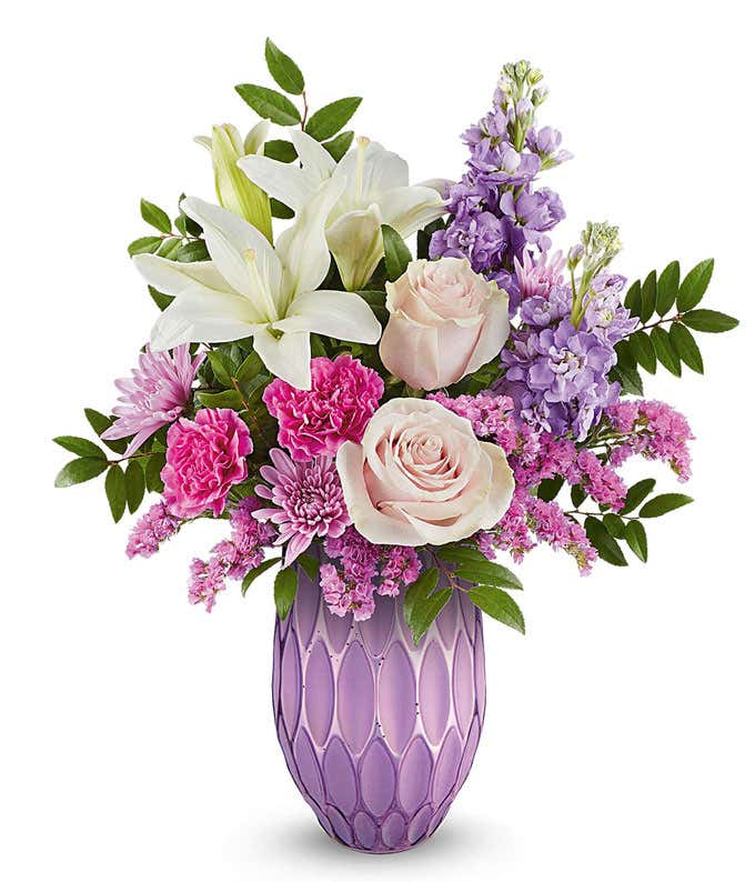 Pink roses, white Asiatic lilies, fuchsia carnations, lavender stock, lavender cushion spray chrysanthemums, raspberry sinuata statice, and floral greenery arranged in a decorative lavender vase with a textured geometric pattern.