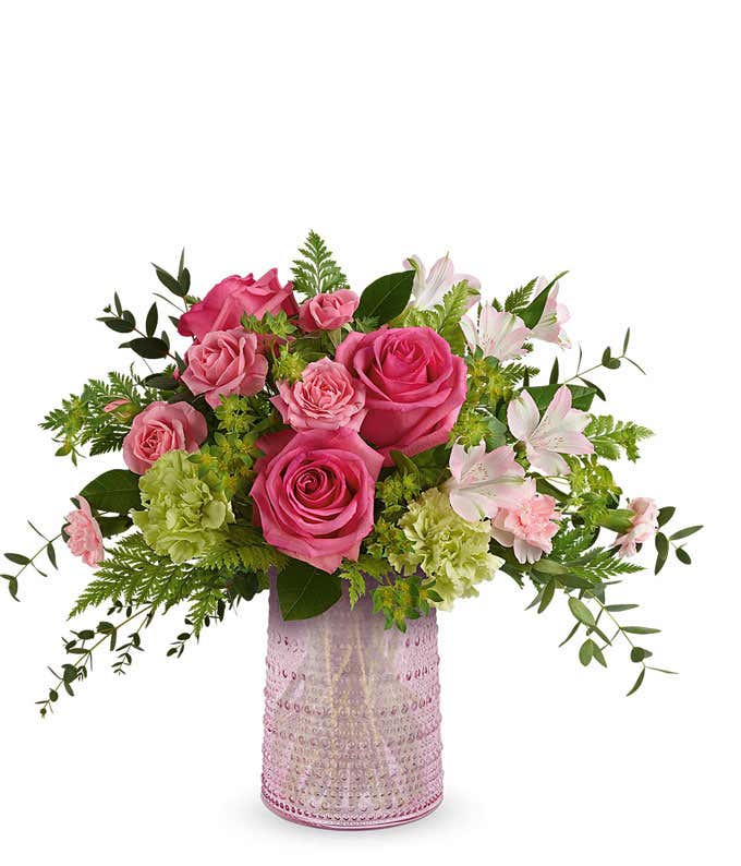 Hot pink roses, pink spray roses, light pink alstroemeria, green carnations, miniature pink carnations, and floral greenery arranged in a decorative pink lace-patterned vase.