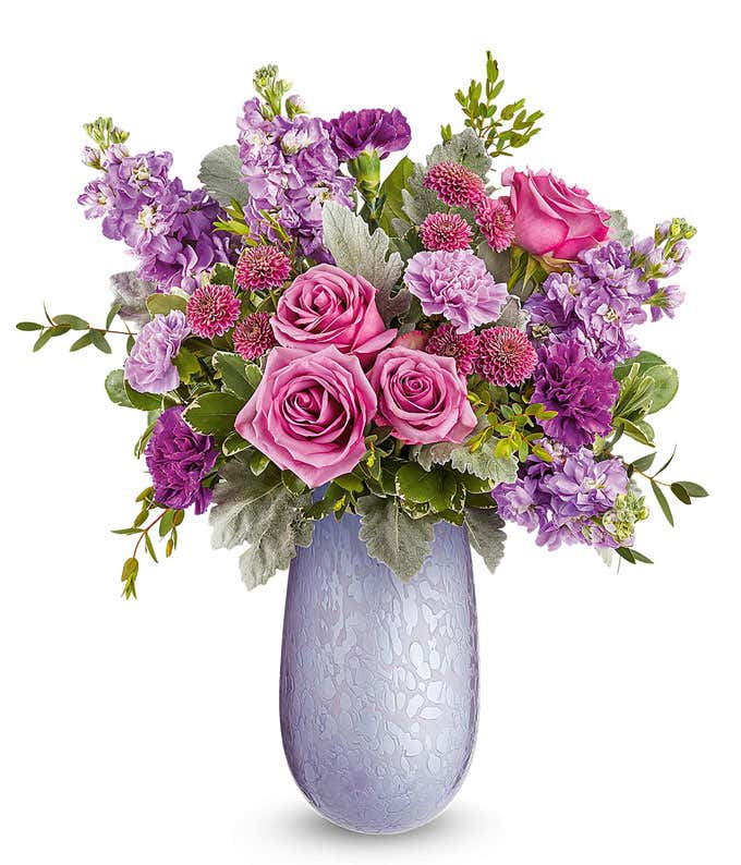 Lavender roses, lavender carnations, purple carnations, lavender stock, purple button spray chrysanthemums, and floral greenery arranged in a decorative lavender glass vase with an embossed floral pattern.