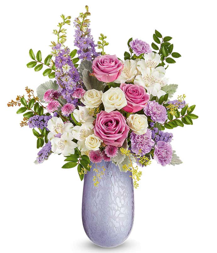 Lavender roses, white spray roses, white alstroemeria, lavender carnations, lavender larkspur, lavender button spray chrysanthemums, lavender sinuata statice, and floral greenery arranged in a decorative lavender glass vase with a crackle pattern.