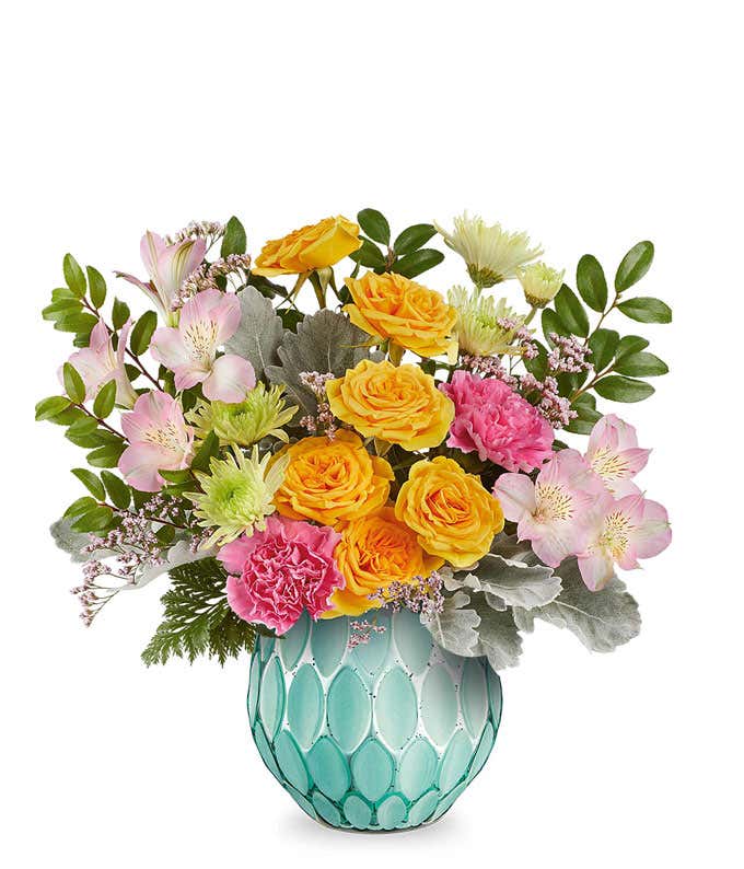 Pink roses, yellow spray roses, hot pink carnations, pink alstroemeria, green button spray chrysanthemums, pink waxflower, and assorted greenery arranged in a textured turquoise glass vase.