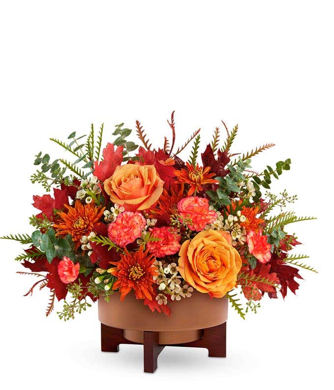 Fall flower arrangement with peach roses, orange carnations, rust-colored daisy chrysanthemums, white waxflower, and assorted autumn leaves in a brown ceramic container on a wooden stand.