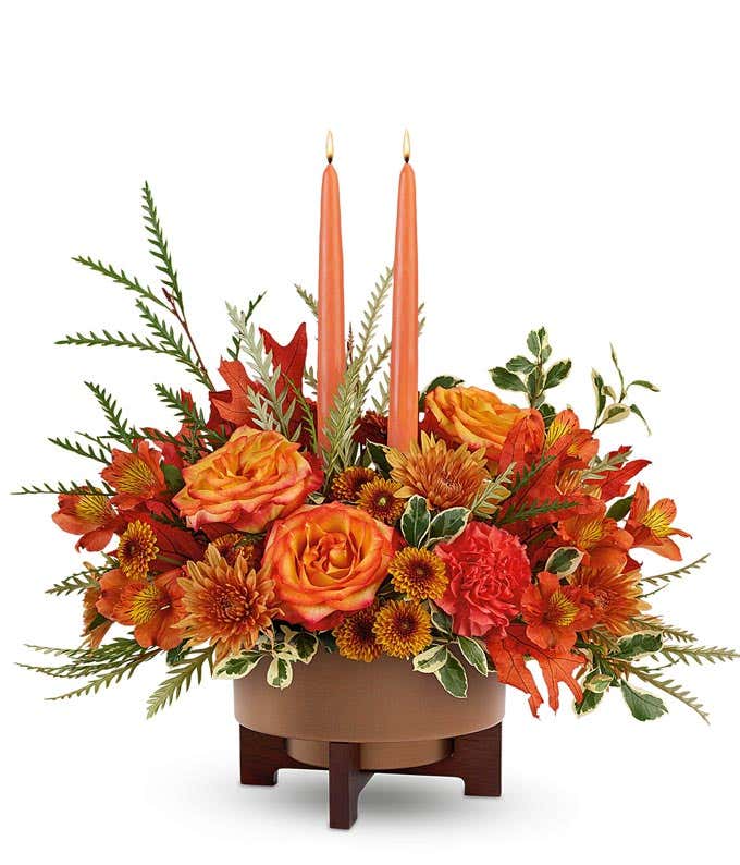Fall centerpiece with orange roses, orange spray roses, red carnations, bronze cushion spray chrysanthemums, fall leaves, and greenery, arranged around two orange taper candles in a brown ceramic planter with wooden stand.