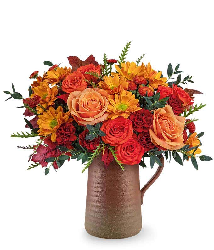 Fall flower arrangement with orange roses, orange spray roses, maroon carnations, brown button spray chrysanthemums, and autumn leaves in a brown ceramic pitcher.