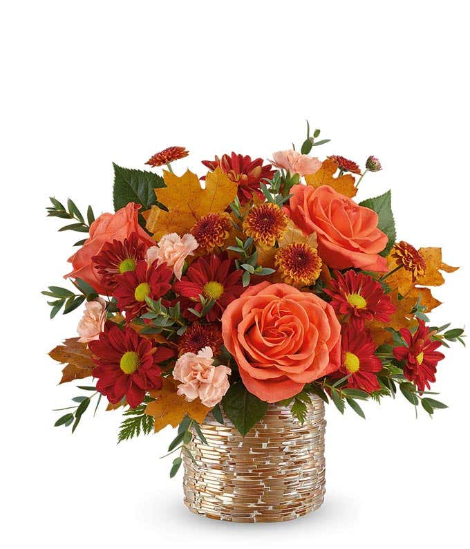 Fall floral arrangement with orange roses, red daisy poms with yellow centers, rust chrysanthemums, light peach carnations, and autumn leaves in a textured gold-glass vase.