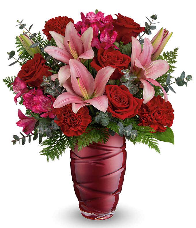 A romantic Valentine's Day bouquet featuring pink lilies, red roses, red carnations, dark pink alstroemeria, and lush greenery, arranged in a deep red swirl-patterned glass vase.