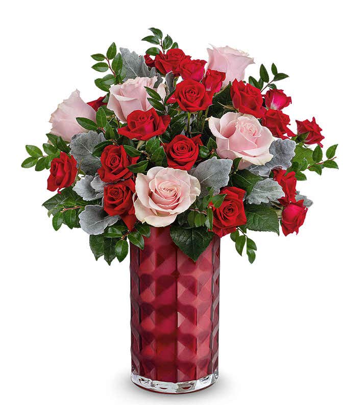 A Valentine's Day bouquet featuring pink roses, red roses, red spray roses, dusty miller, and lush greenery, arranged in a faceted deep red glass vase.