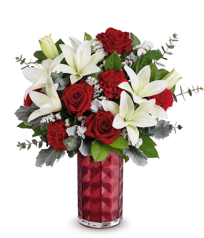 A Valentine's Day bouquet featuring white lilies, red roses, red carnations, white filler flowers, dusty miller, eucalyptus, and lush greenery, arranged in a faceted deep red glass vase.