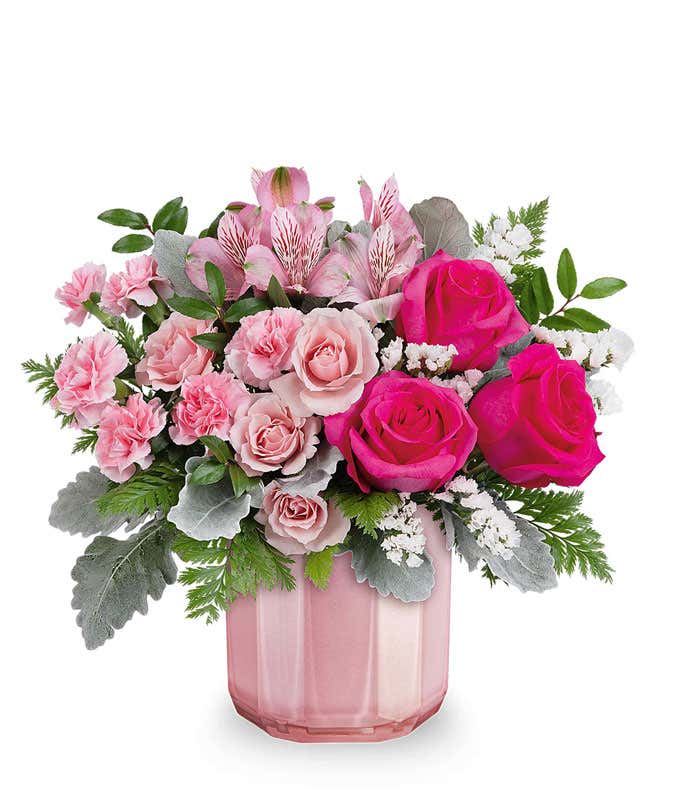 A Valentine's Day bouquet featuring hot pink roses, light pink spray roses, pink alstroemeria, light pink carnations, white filler flowers, dusty miller, and lush greenery, arranged in a soft pink ribbed glass vase.