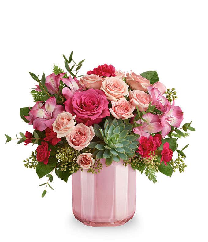 A Valentine's Day bouquet featuring hot pink roses (deluxe and premium sizes only), light pink spray roses, pink alstroemeria, dark pink carnations, a fresh green succulent, seeded eucalyptus, and lush greenery, arranged in a soft pink ribbed glass vase