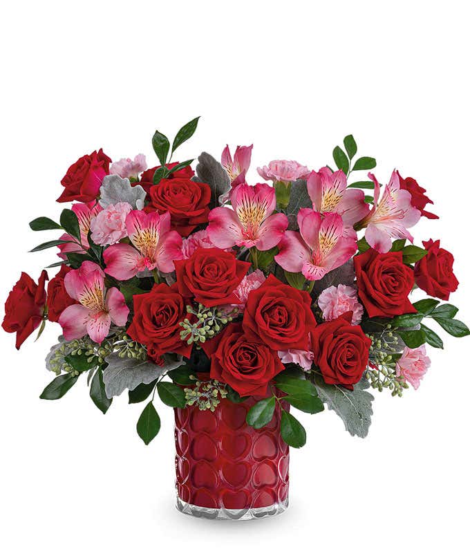 A Valentine's Day bouquet featuring red roses, light pink roses (available in premium size only), pink alstroemeria, pink carnations, seeded eucalyptus, dusty miller, and lush greenery, arranged in a bold red textured glass vase.