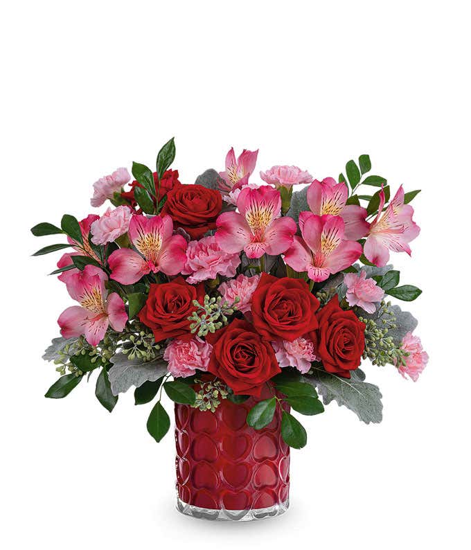 A Valentine's Day bouquet featuring red roses, light pink roses (available in premium size only), pink alstroemeria, pink carnations, seeded eucalyptus, dusty miller, and lush greenery, arranged in a bold red textured glass vase.