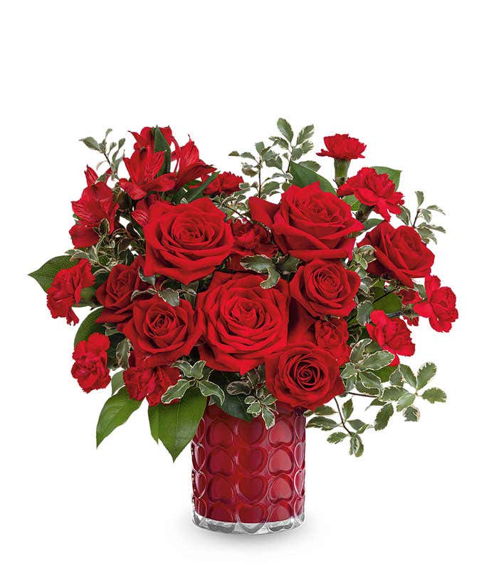 A Valentine's Day bouquet featuring red roses, red spray roses, red carnations, red alstroemeria, variegated greenery, and lush foliage, arranged in a red glass vase with a textured heart pattern.