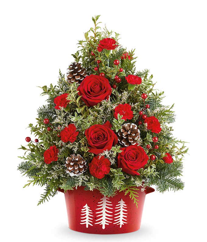 Christmas tree-shaped floral arrangement with red roses, red carnations, pinecones, berries, and greenery in a red container decorated with white tree silhouettes.
